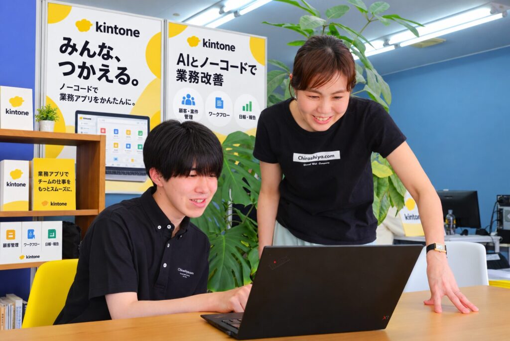 kintoneのポスターの前で、ノートパソコンを操作するスタッフと横からサポートするスタッフ